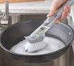 Hand using a scrubbing brush to clean a soapy pan in a kitchen sink.