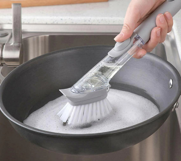 Hand using a scrubbing brush to clean a soapy pan in a kitchen sink.