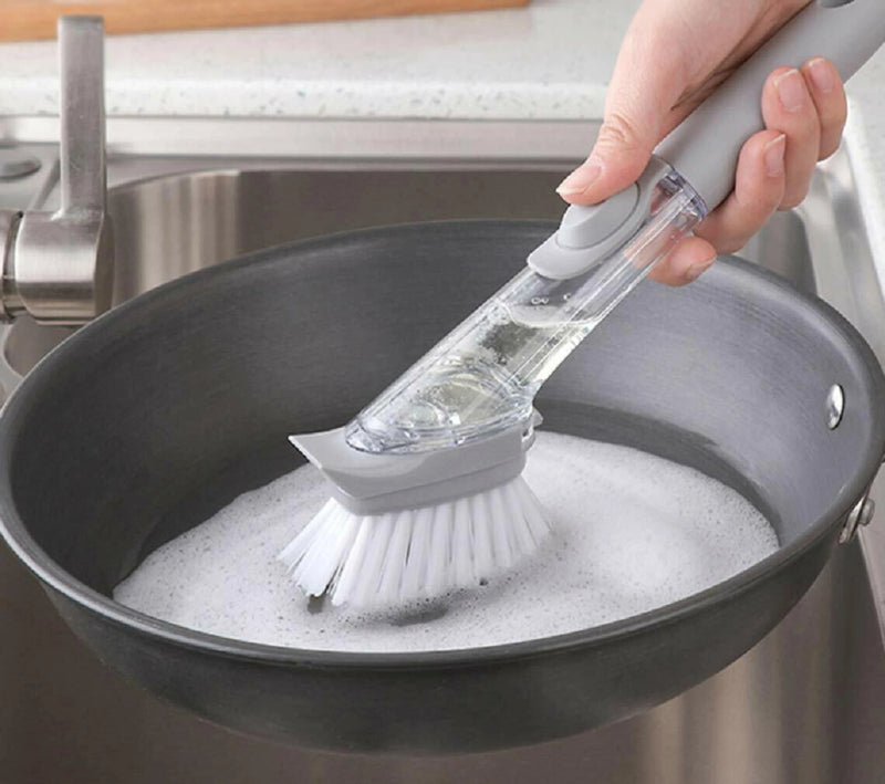 Hand using a scrubbing brush to clean a soapy pan in a kitchen sink.