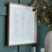 Solli feather art hung on a dark green wall beside a vase of foliage